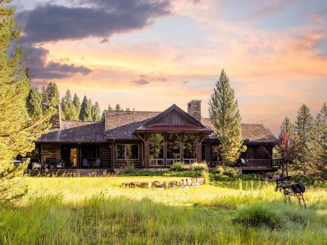 Una casa grande de madera se alza junto a un campo de hierba bajo un cielo colorido al atardecer.