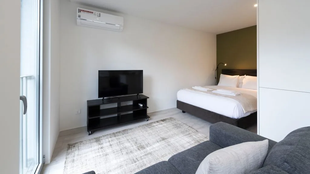 A bedroom with a bed, television, and gray seating beside a large window.