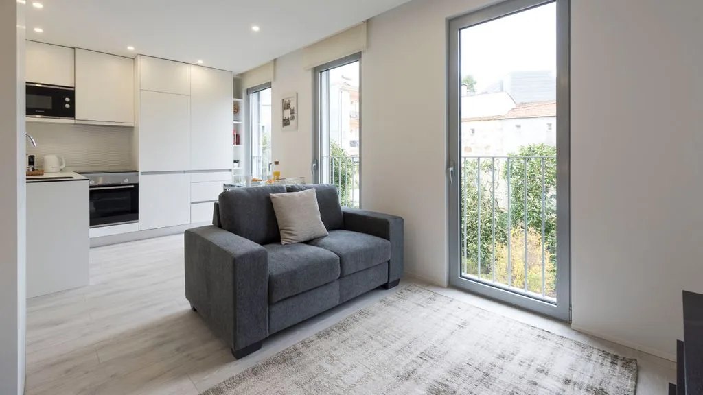 A bright living area with gray seating beside large windows and an open kitchen.