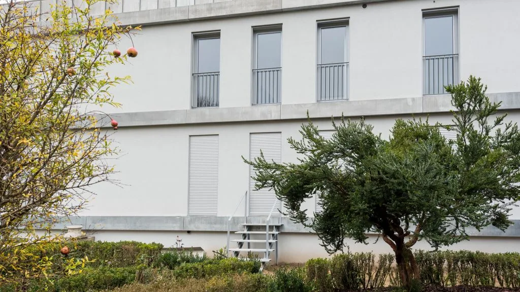 A white building stands behind a landscaped garden with trees and shrubs.