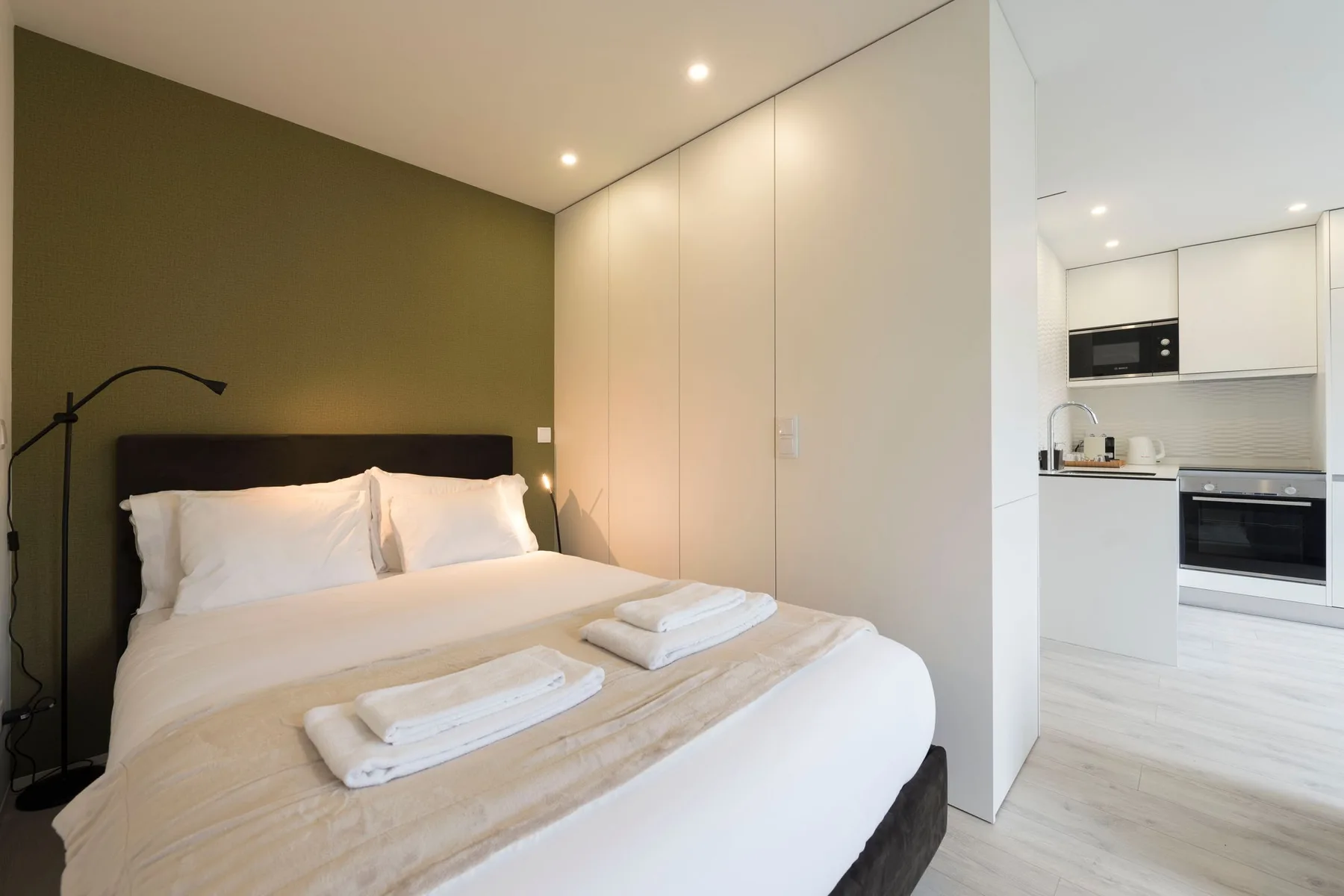 A bedroom with a neatly made bed and a kitchen visible through the open doorway.