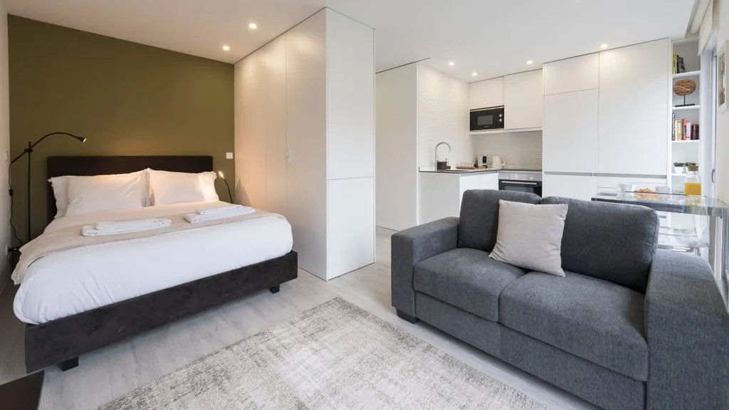 A modern studio room with a bed, seating, and a compact kitchen.