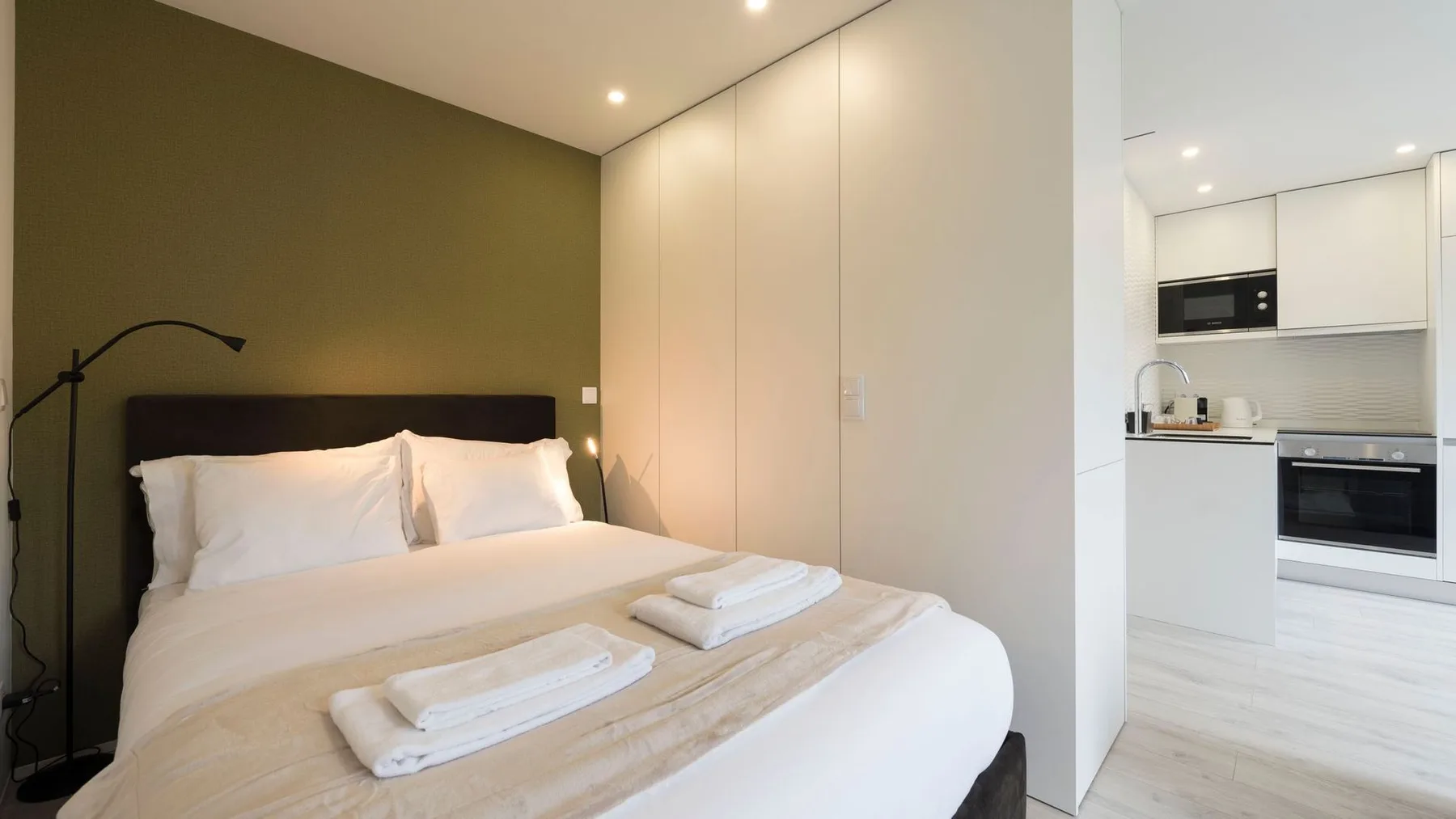A bedroom with a neatly made bed and a kitchen visible beyond the doorway.