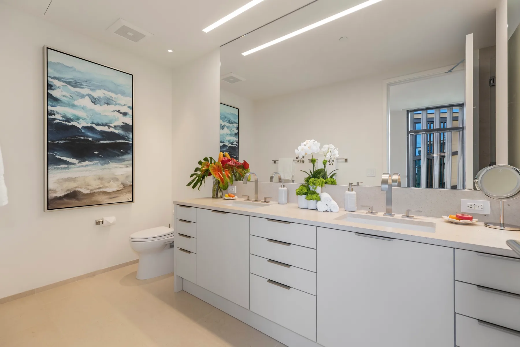 A bright bathroom has a long vanity with a wide mirror and floral arrangements.