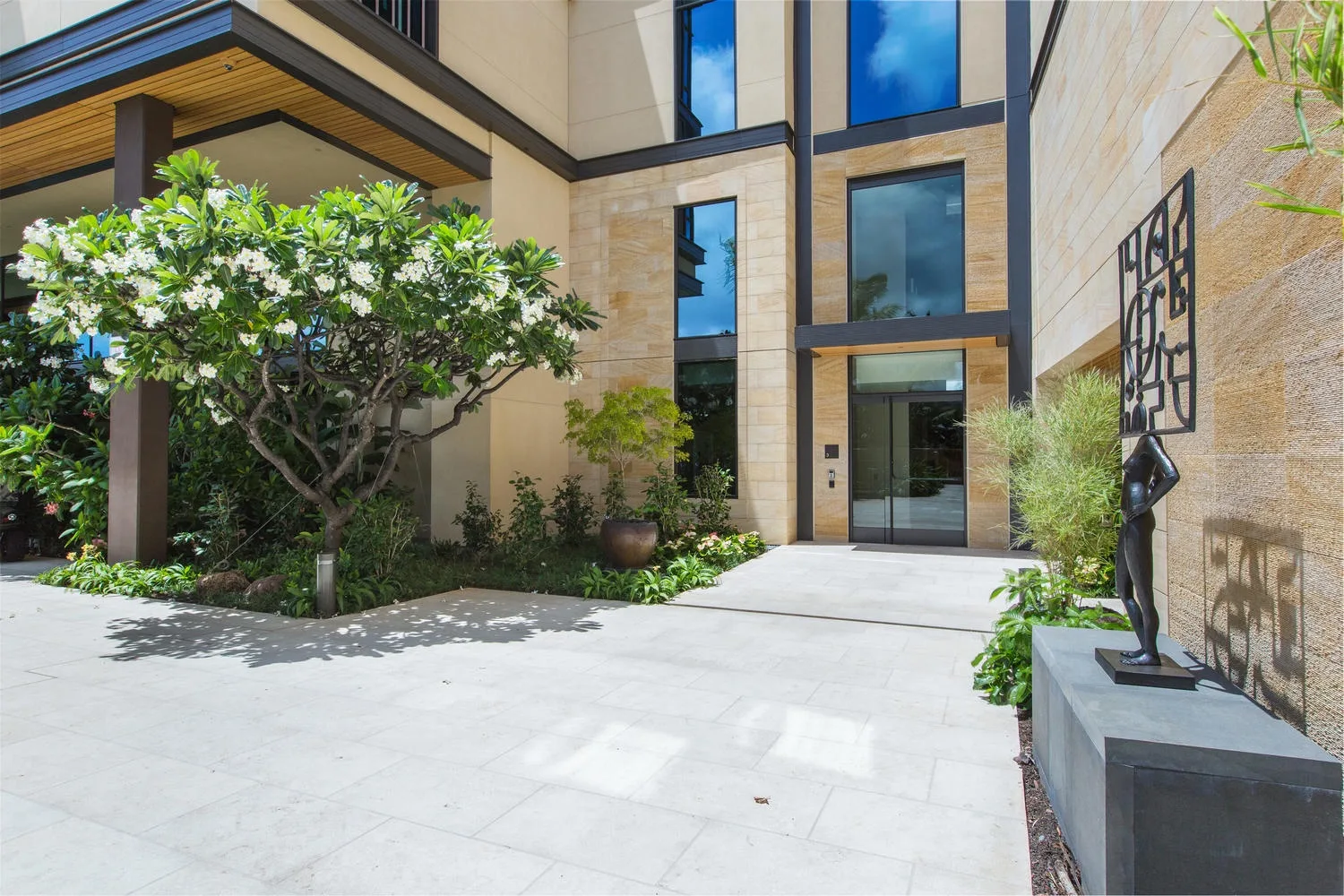 A modern building entrance opens onto a paved courtyard with trees and a sculpture.