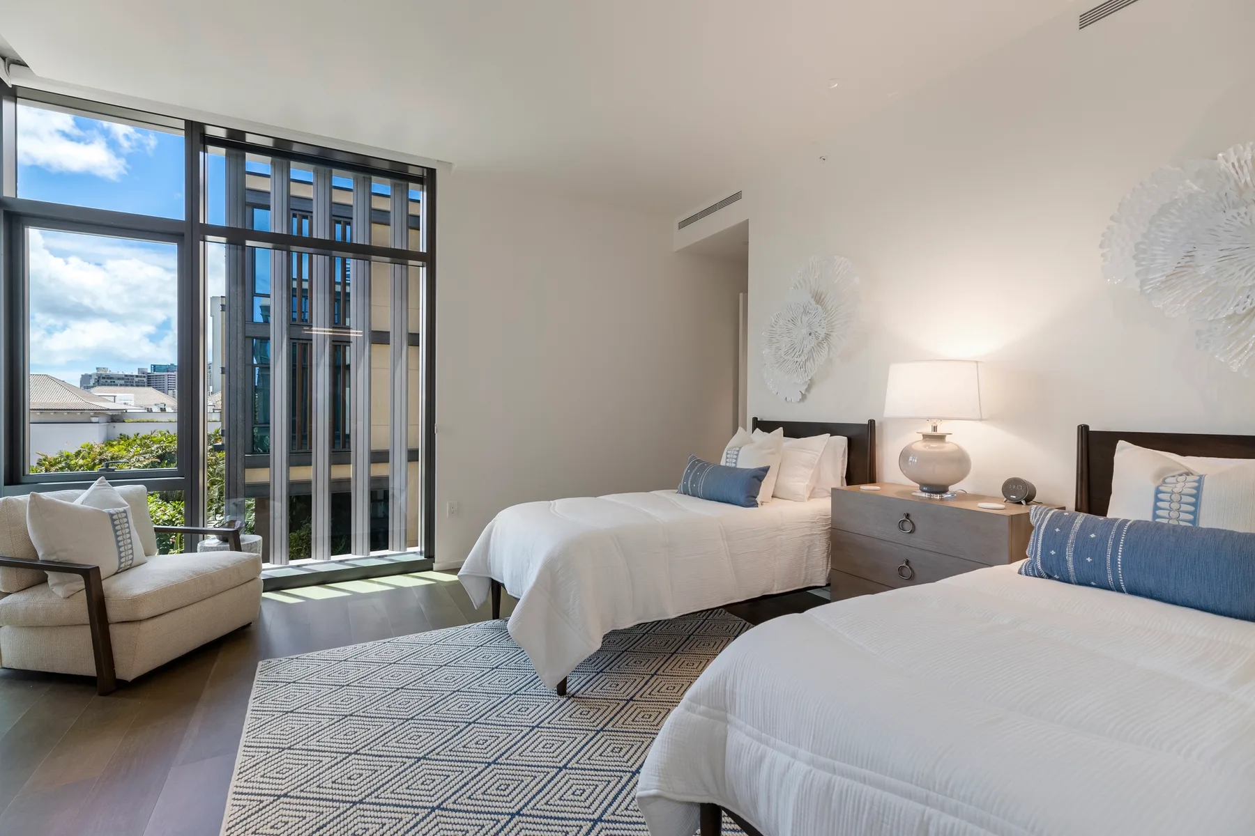 A bright bedroom with two beds and large windows overlooking nearby buildings.