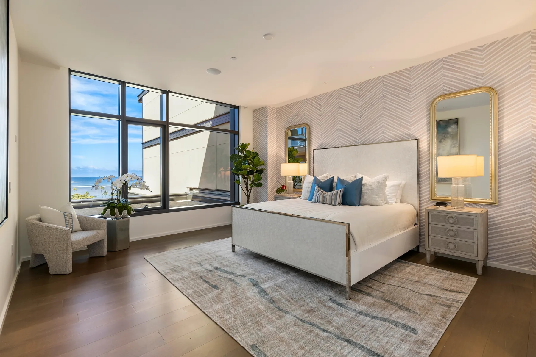 A bright bedroom with a large bed, patterned walls, and windows overlooking the water.