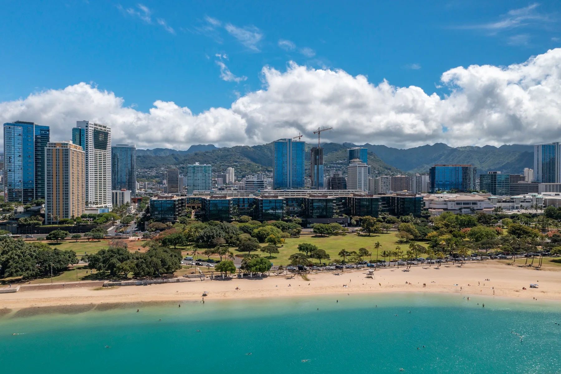A coastal beach and turquoise water border a green park and city skyline.