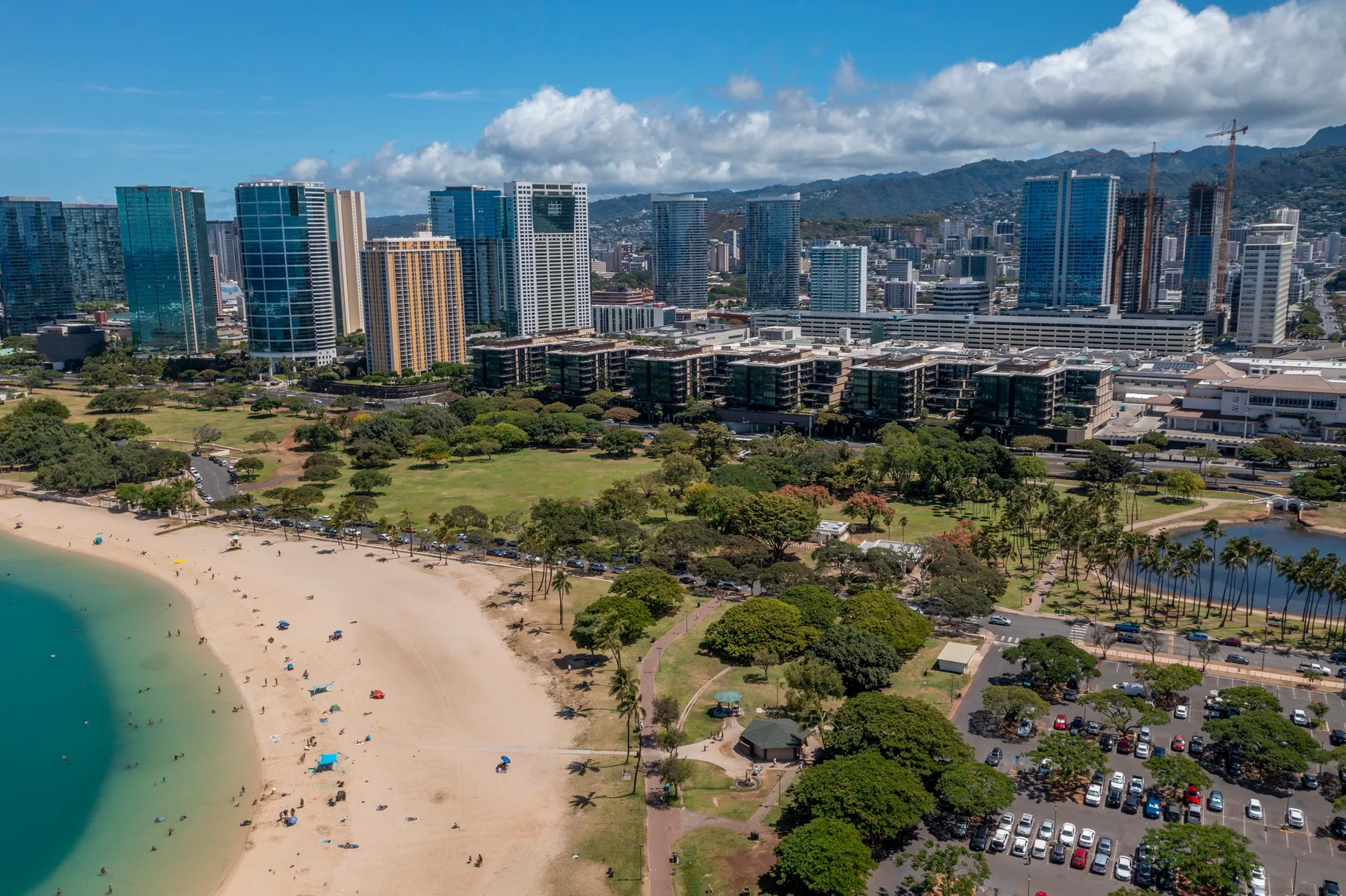 Aerial view of a sandy beach beside a coastal park and city skyline.