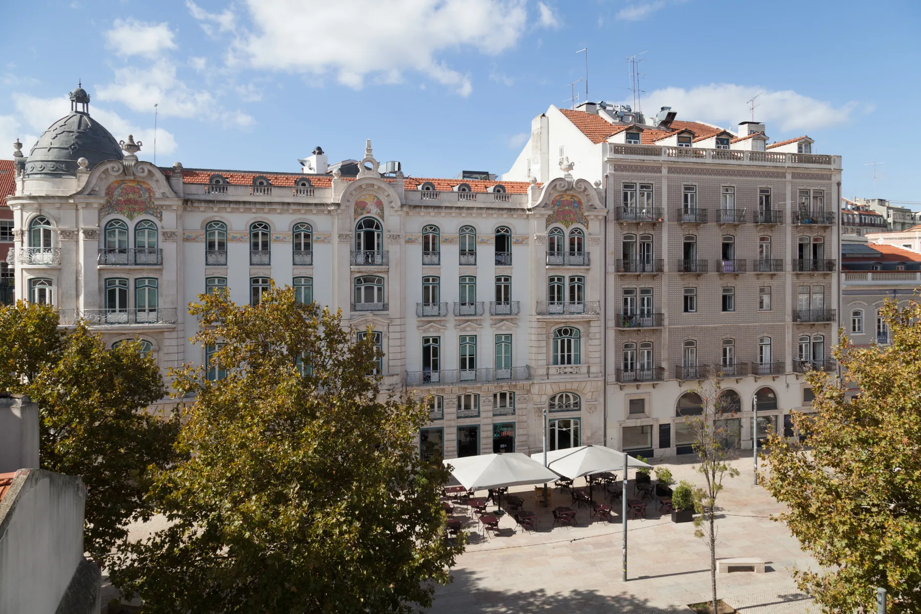 Un edificio urbano ornamentado da a una plaza pavimentada con mesas de café al aire libre.