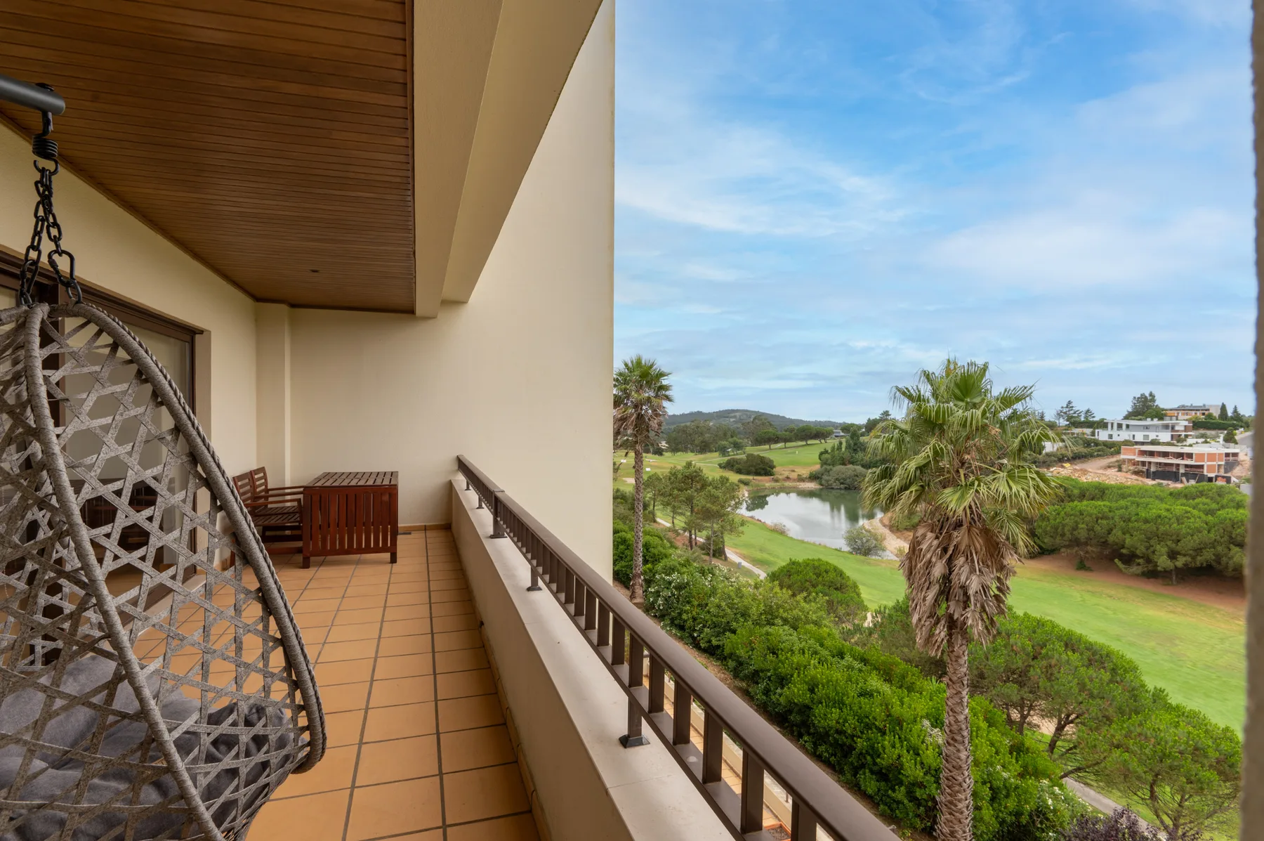 A covered balcony with a hanging seat overlooks palm trees and a green landscape.