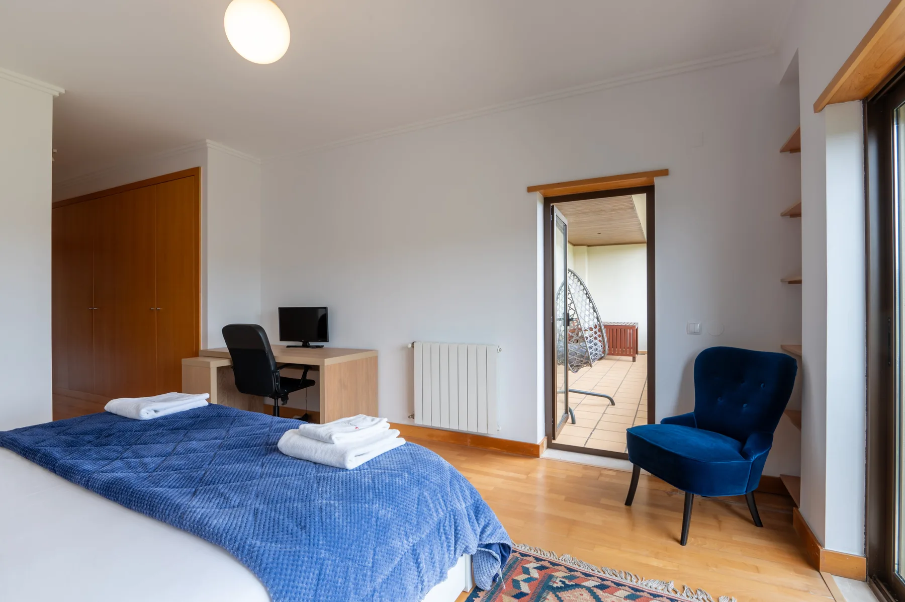 A bedroom with blue bedding, a desk, and a doorway opening onto a balcony.