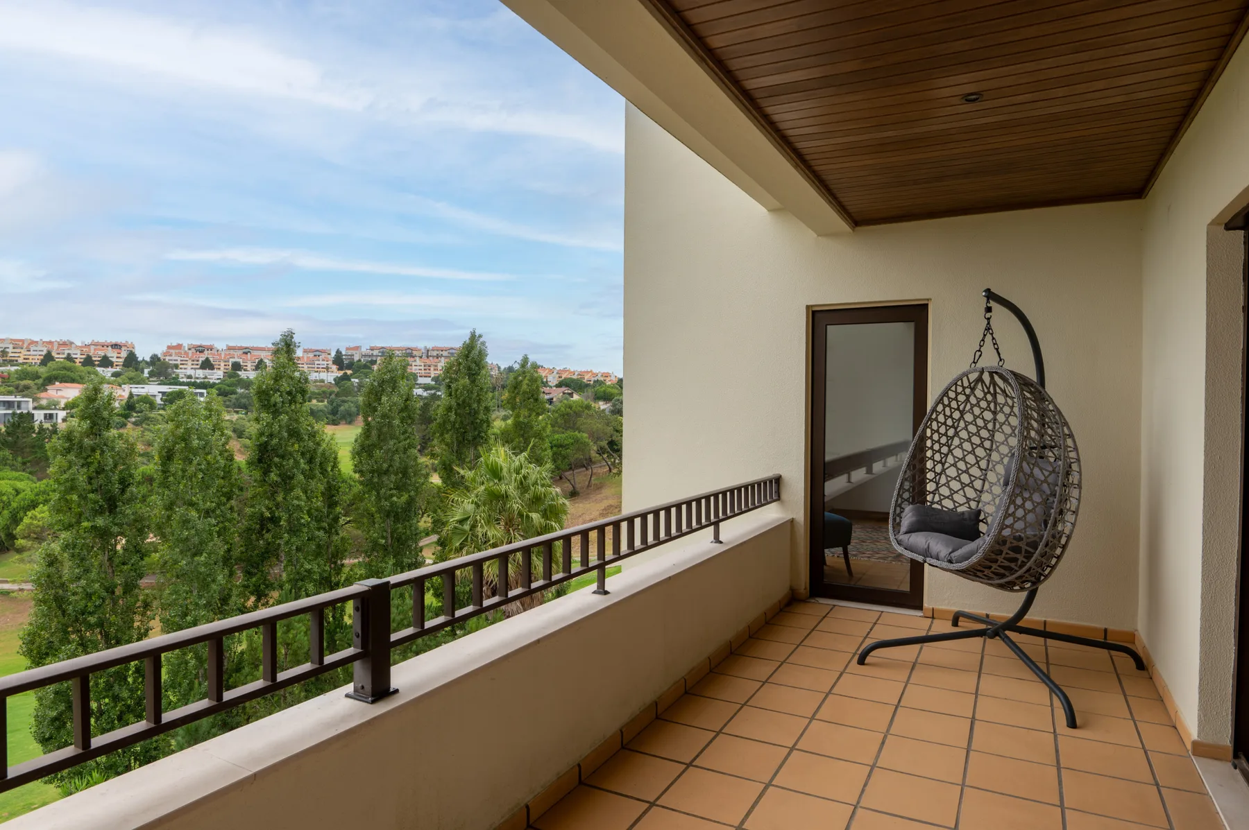 A covered balcony with a hanging seat overlooks trees and buildings.
