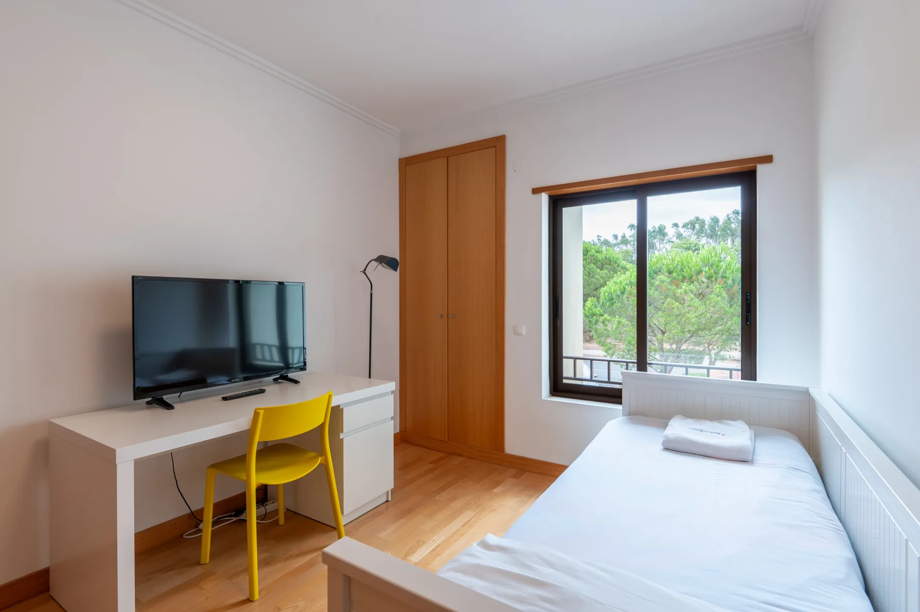 A bedroom with a bed, desk, and window overlooking greenery.