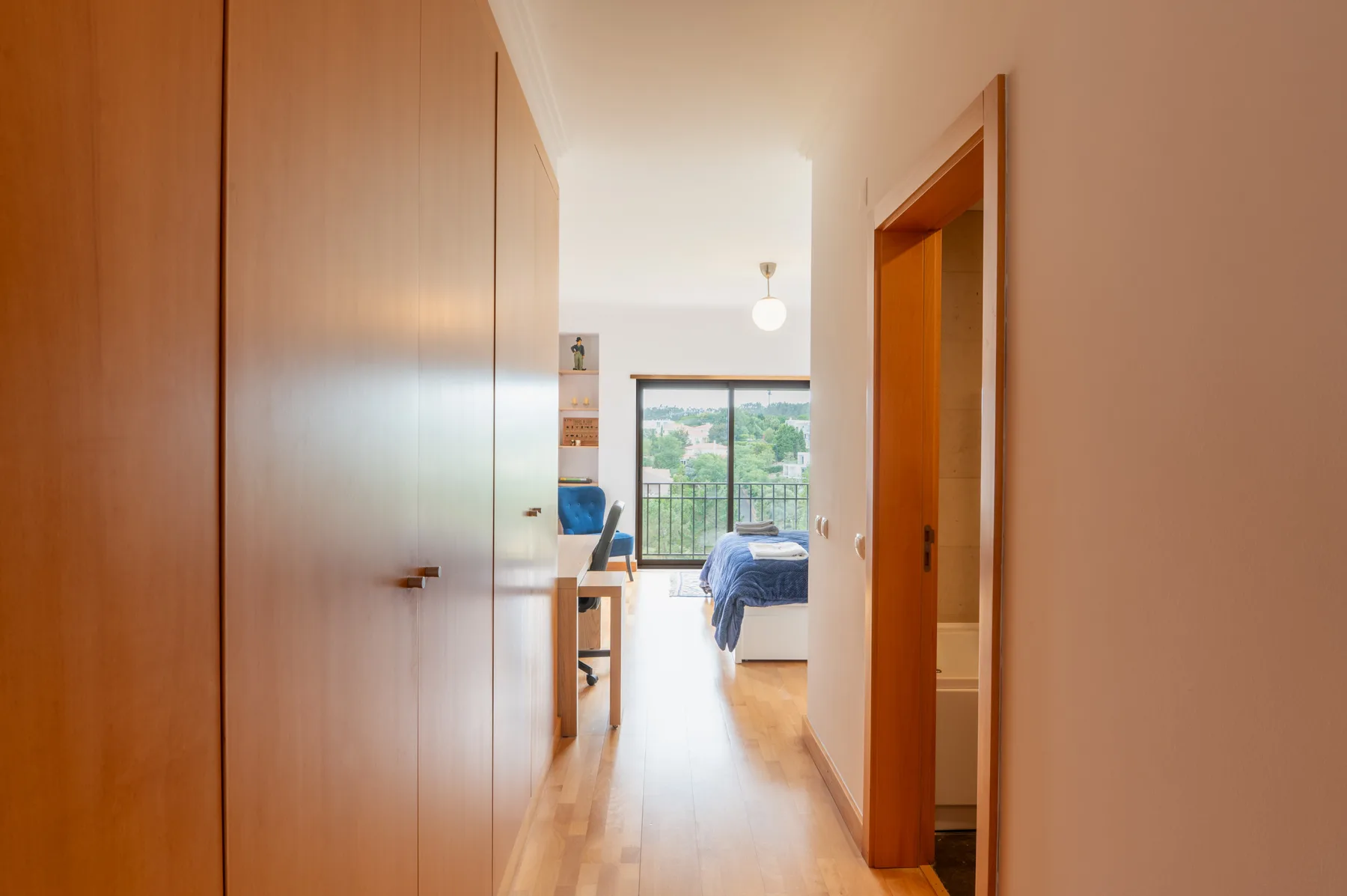 A hallway opens toward a bedroom with a bed and large windows.