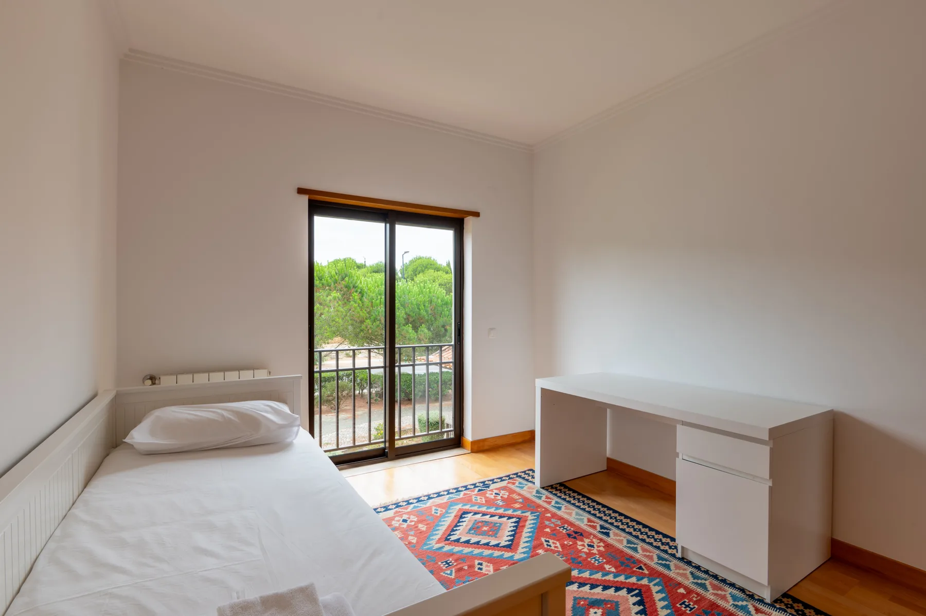 A bedroom with a single bed, a desk, and a balcony door overlooking greenery.