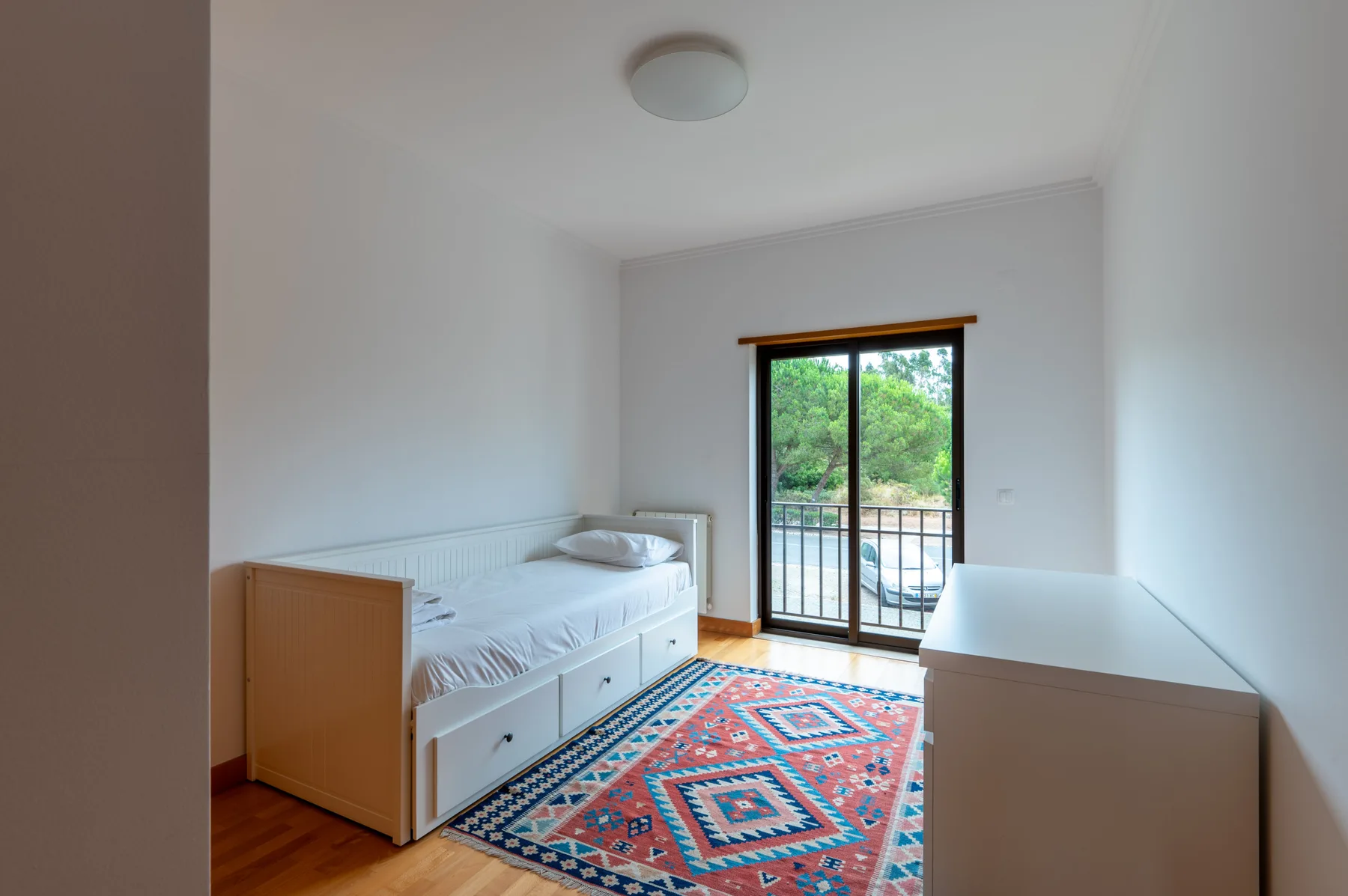 A bright bedroom with a single bed, a colorful rug, and glass doors opening to a balcony.