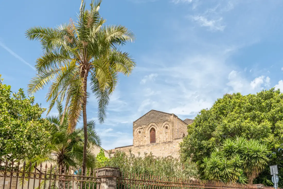 Un edificio de piedra se alza tras árboles frondosos y una palmera alta bajo el cielo azul.