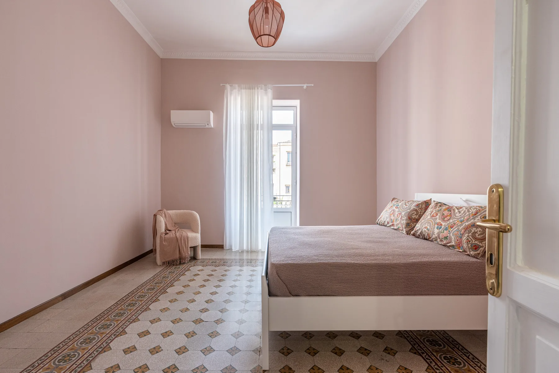 A bedroom with a bed, patterned pillows, and a tall window with sheer curtains.