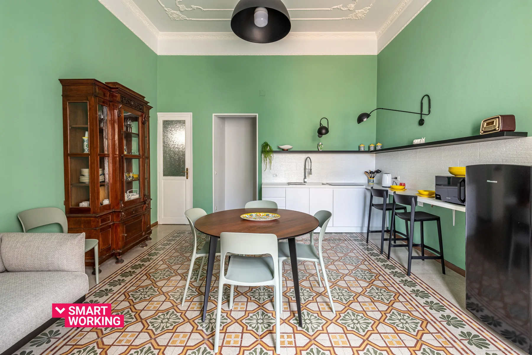 Zona de comedor y cocina con una mesa redonda, paredes verdes y suelo de baldosas estampadas.