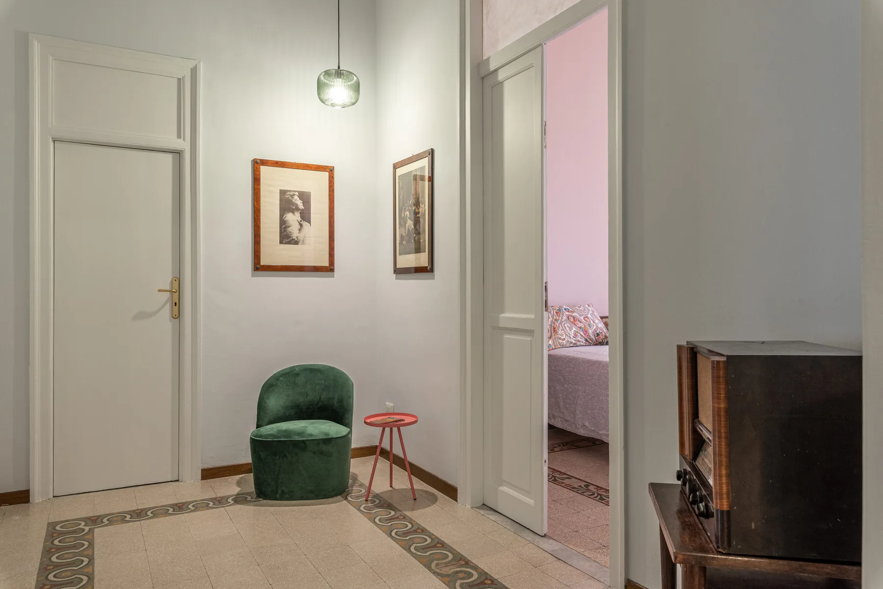 Una pequeña zona de estar con asiento verde y cuadros junto a la entrada abierta de un dormitorio.