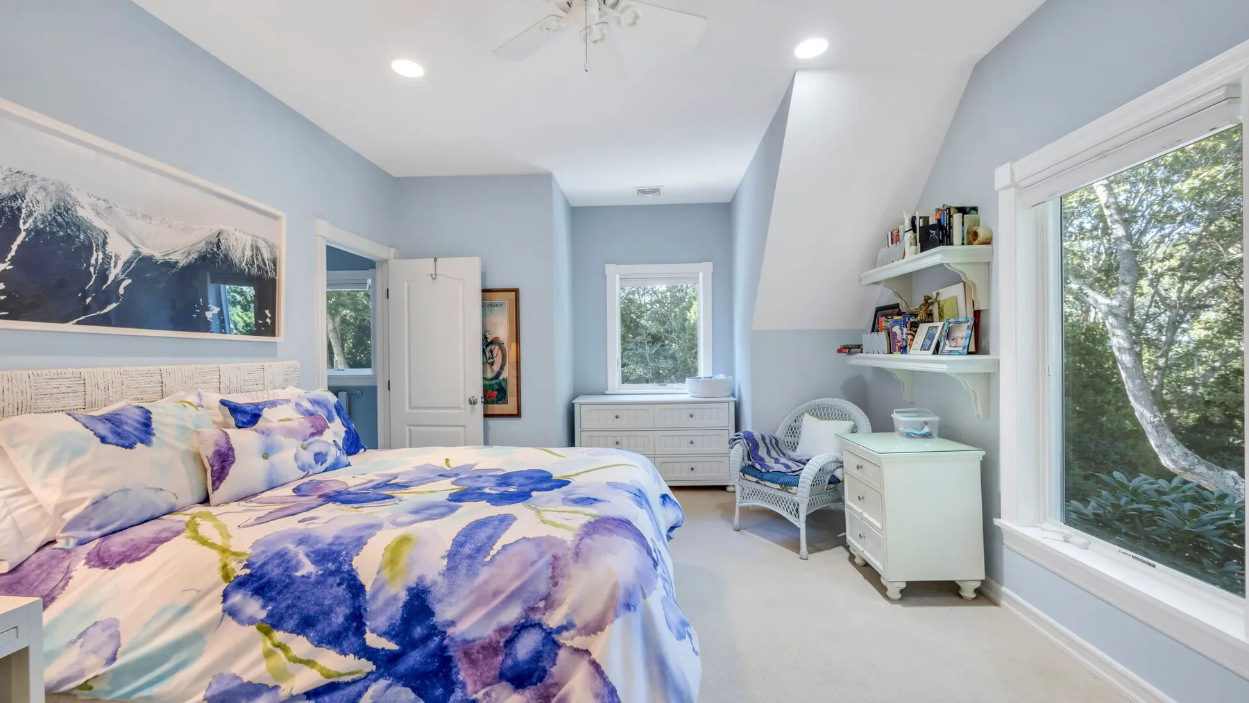 Un dormitorio luminoso con ropa de cama floral, una cómoda y grandes ventanas.