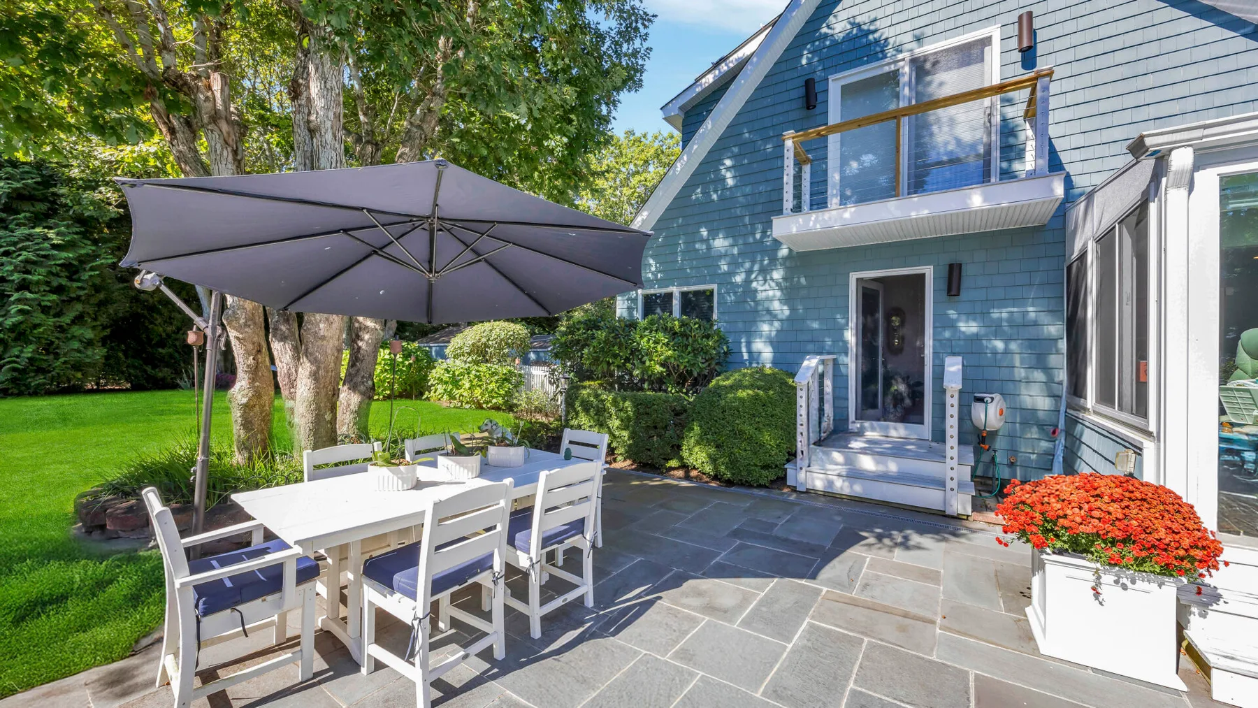 Un patio pavimentado junto a una casa azul tiene asientos al aire libre bajo una sombrilla grande.