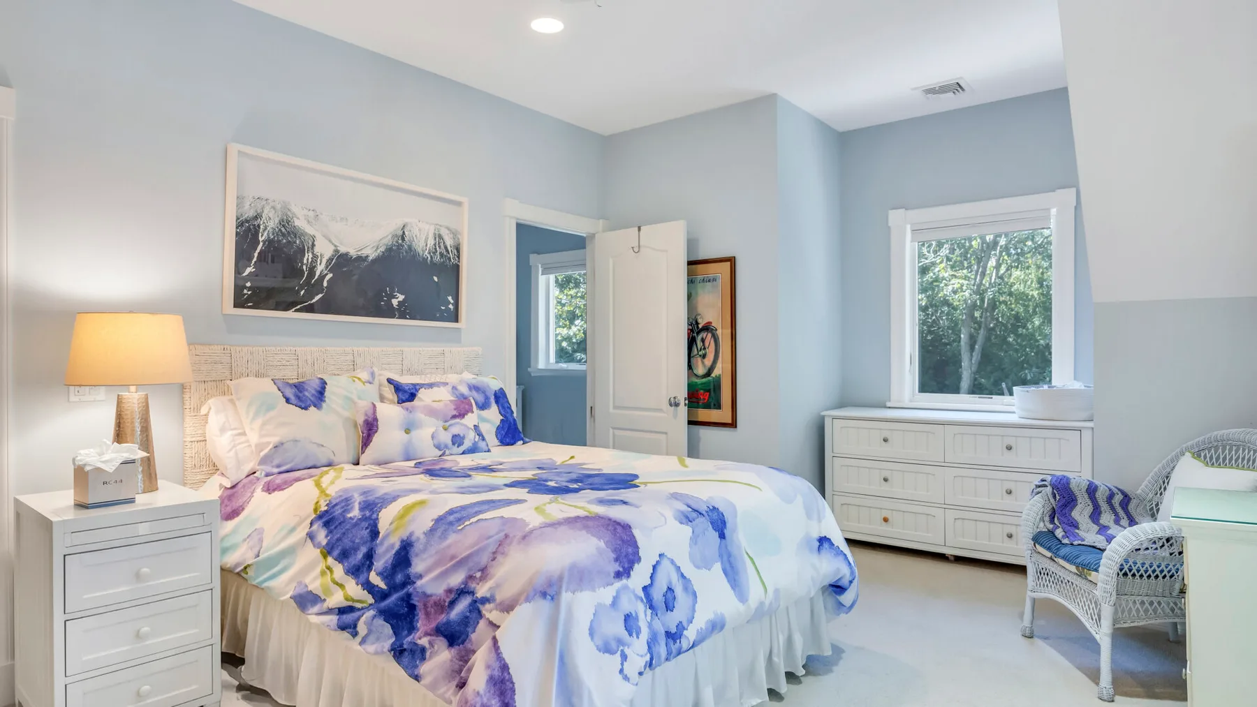 Un dormitorio luminoso con una cama vestida con ropa de cama floral azul y morada.