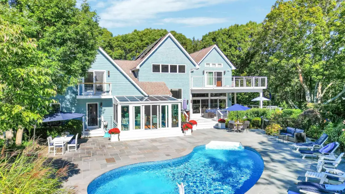 Una casa grande da a una piscina y un patio pavimentado entre árboles frondosos.