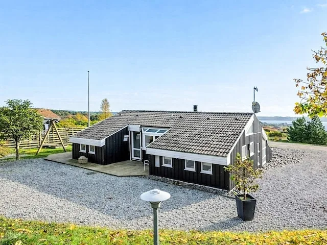 A dark wooden holiday house stands beside a gravel drive under a clear sky.