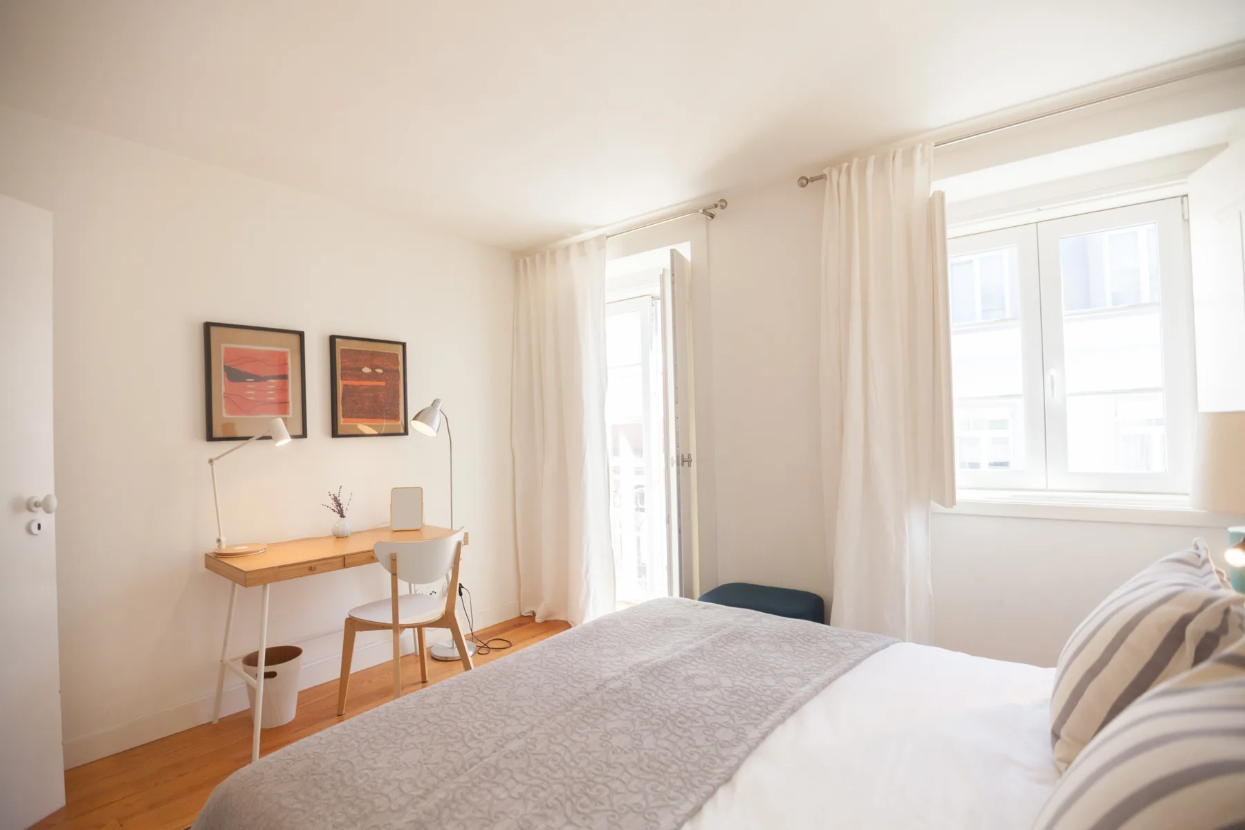 Un dormitorio luminoso con una cama, un escritorio pequeño y ventanas altas.