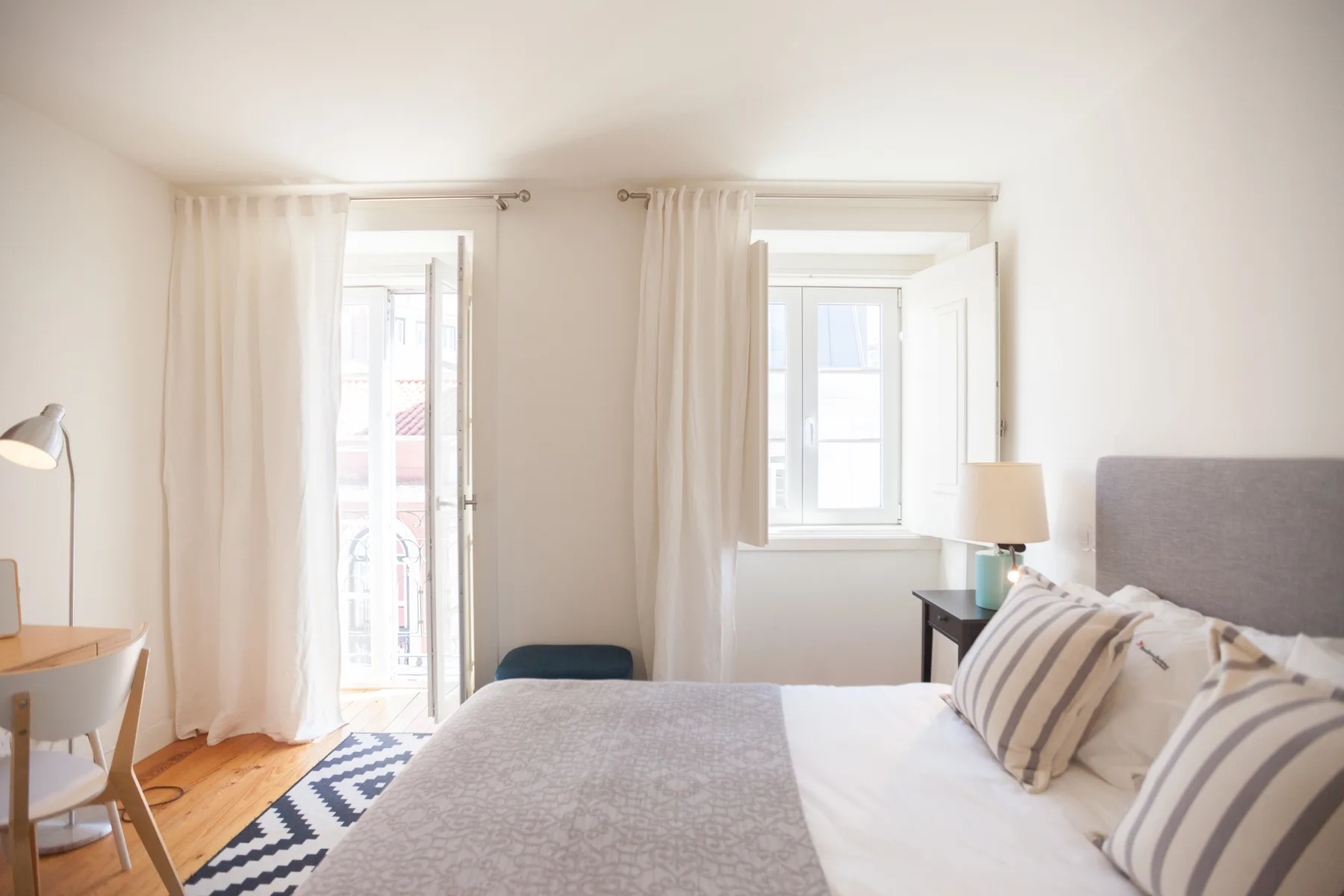 Un dormitorio luminoso con una cama junto a ventanas abiertas y cortinas claras.