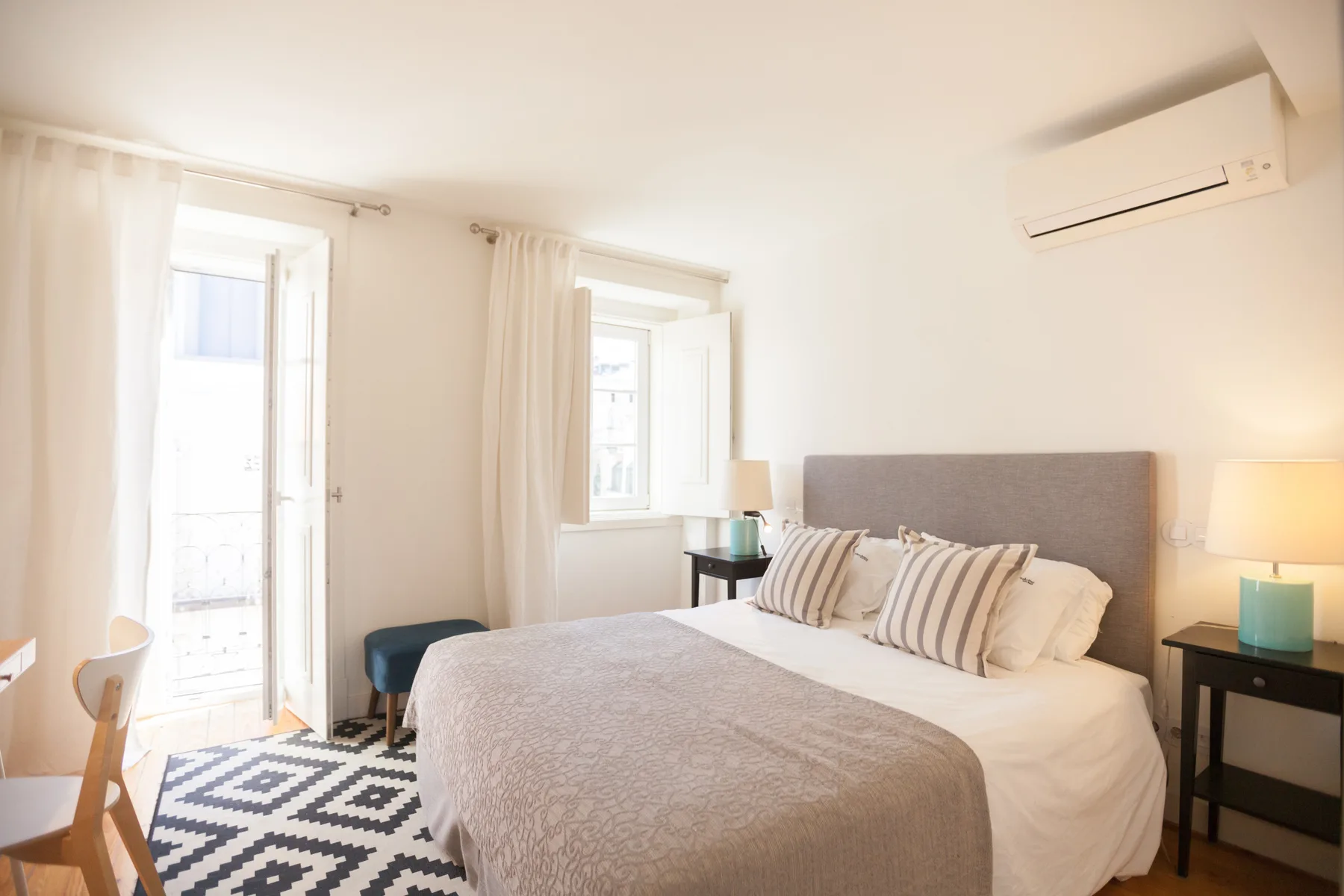 Un dormitorio luminoso con una cama bien hecha y ventanas altas.
