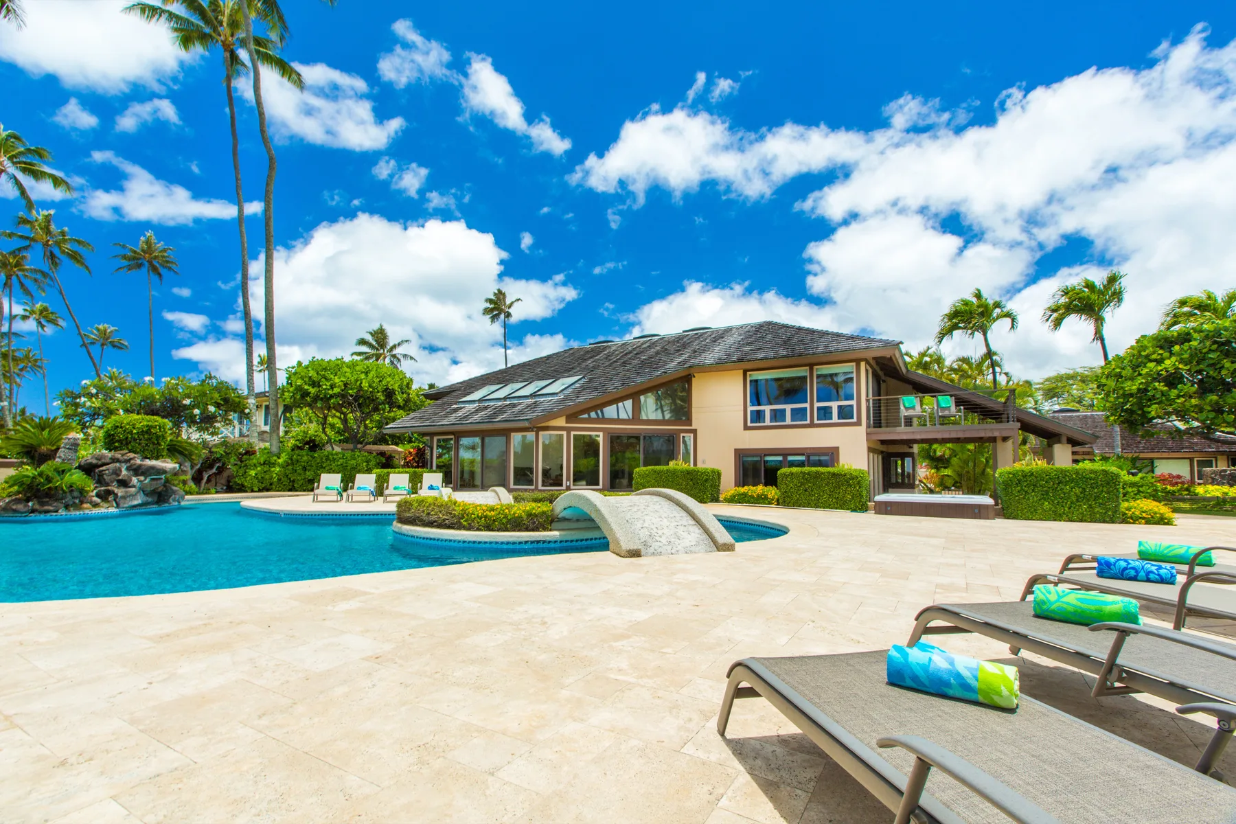 Una zona de piscina tropical con asientos reclinables frente a una casa grande.