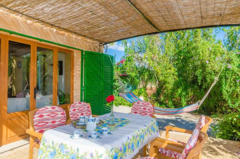Un patio exterior a la sombra con una mesa de comedor y una hamaca colorida entre la vegetación.