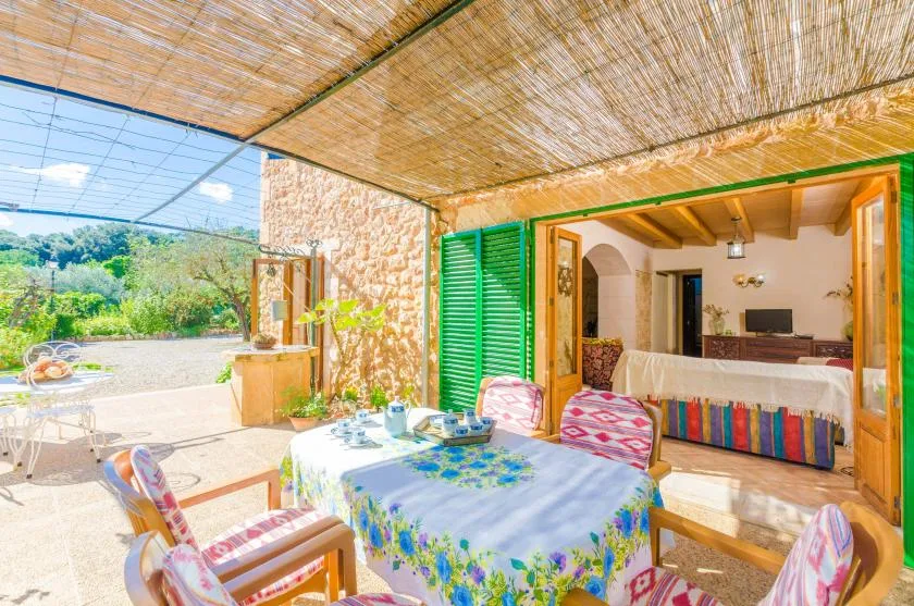 Comedor exterior a la sombra junto a una habitación abierta, con un mantel estampado.