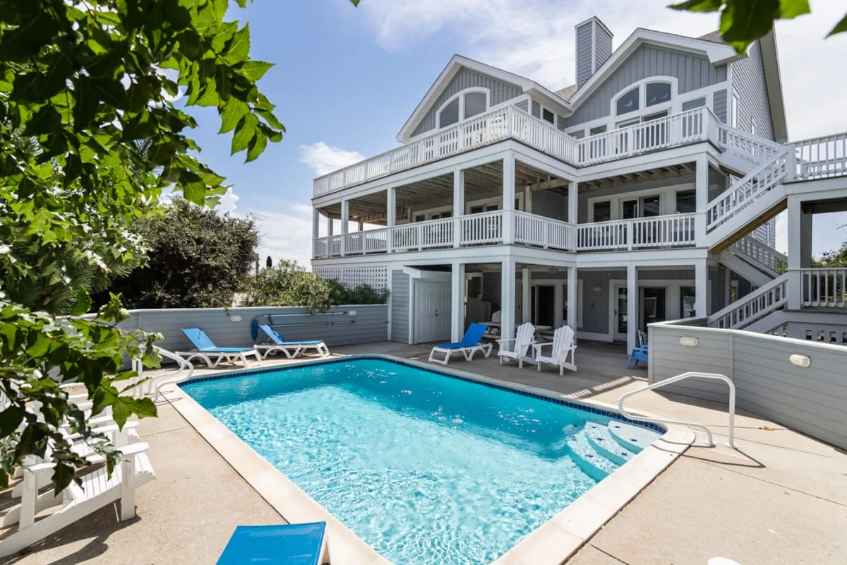 An outdoor swimming pool sits beside a large house with wraparound balconies.
