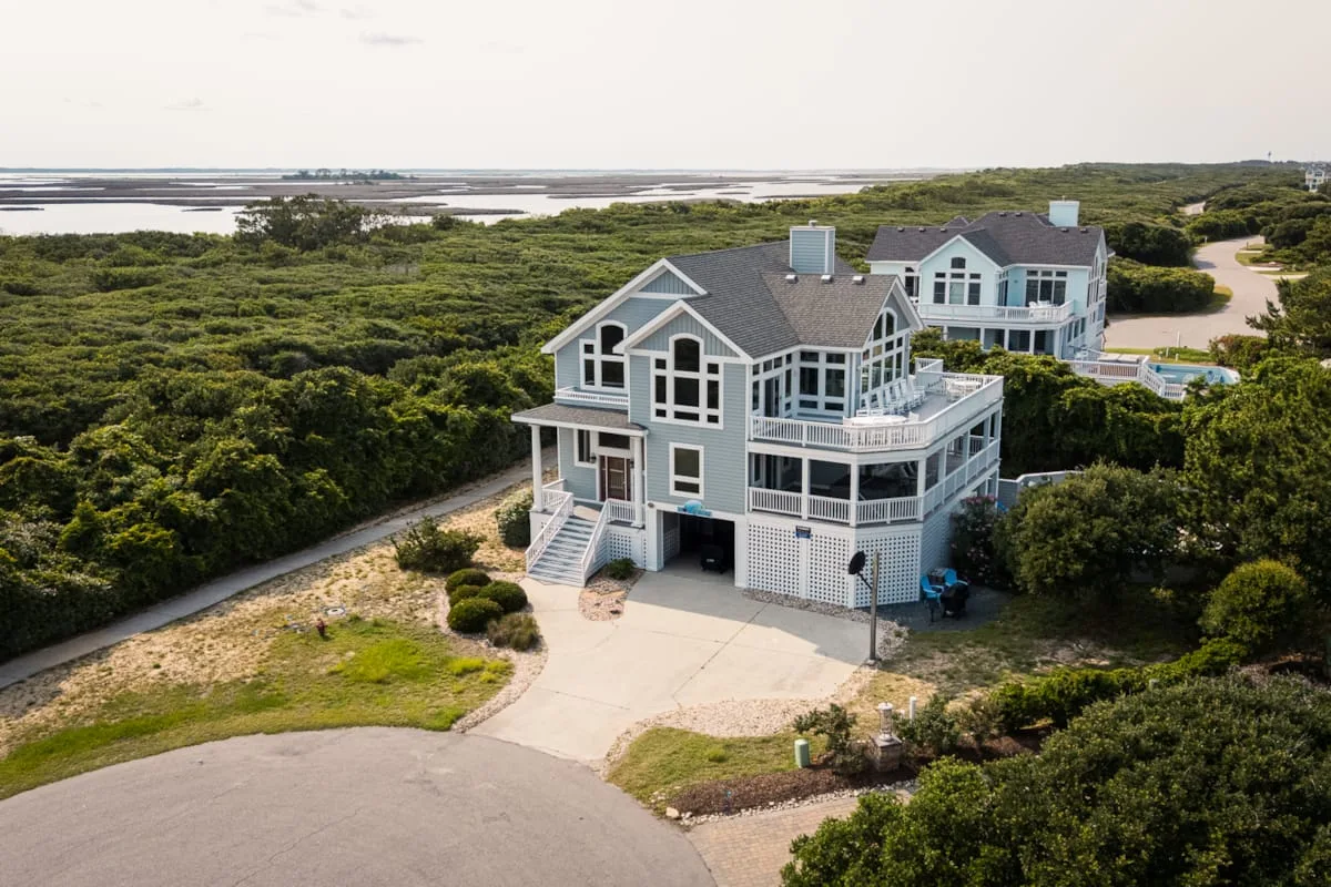 Aerial view of two coastal houses surrounded by dense greenery, with water in the distance.