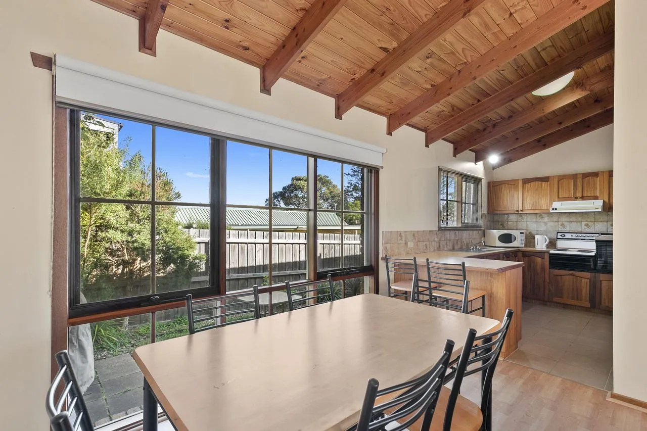 Comedor junto a grandes ventanales, con cocina abierta y vigas de madera a la vista.