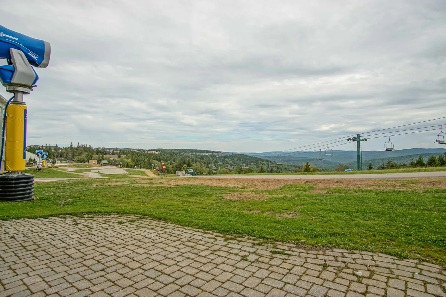 An open grassy landscape with paved paths and ski lift cables under a cloudy sky.