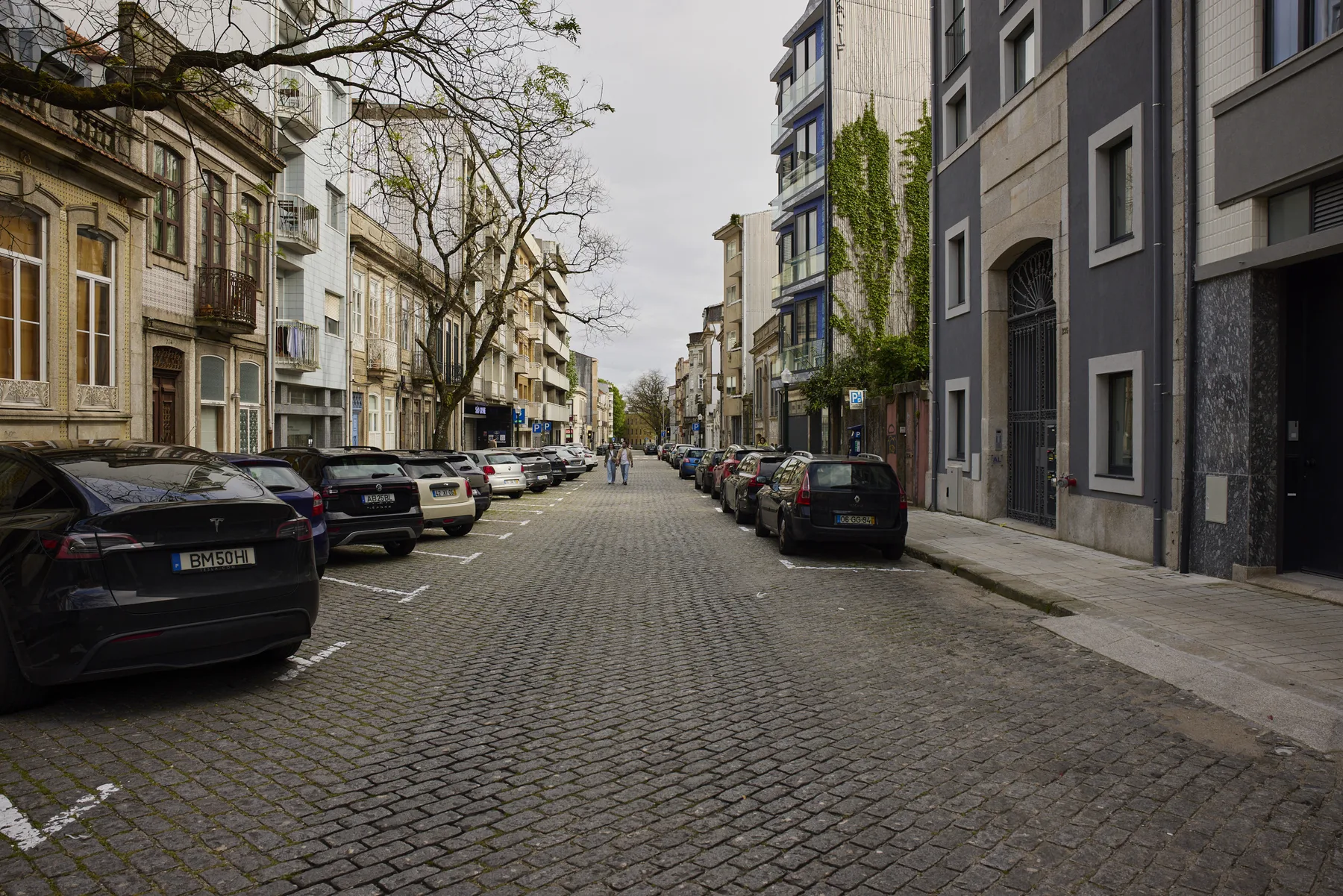 A cobblestone city street lined with parked cars and multi-story buildings.