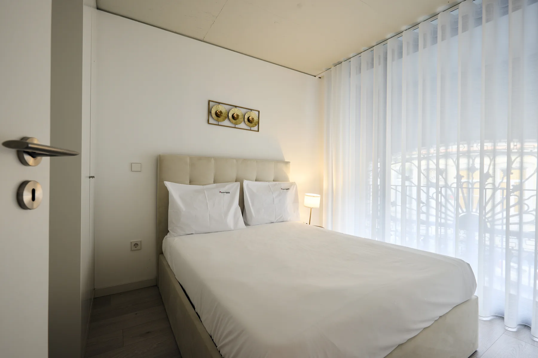 A bedroom with a bed beside large windows covered by sheer curtains.