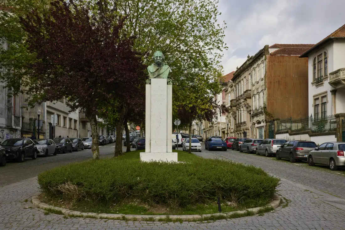 A tree-lined urban street surrounds a grassy island with a bust on a pedestal.