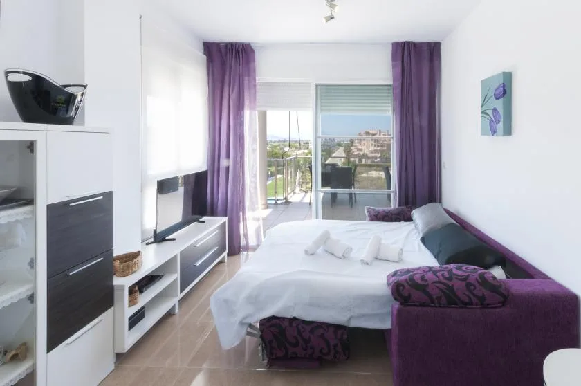 A bright bedroom with a bed beside a purple sofa and balcony doors.