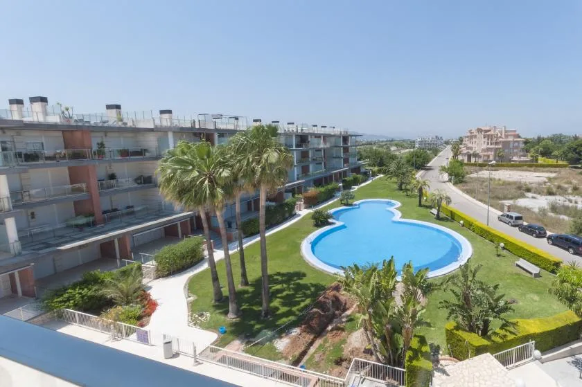 A residential complex surrounds a curved swimming pool, with palm trees and landscaped lawns.