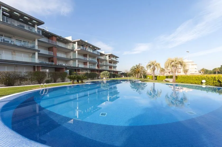 A large outdoor swimming pool sits beside a modern apartment building and landscaped grounds.