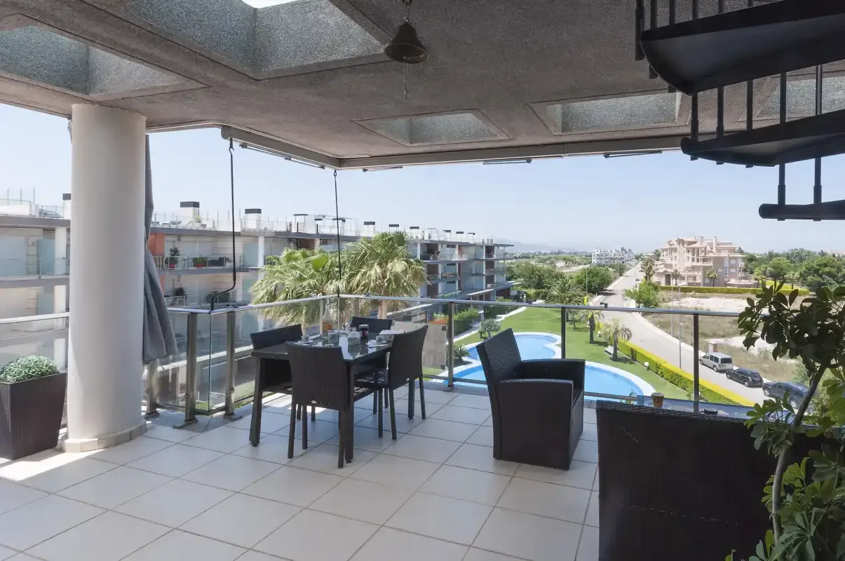 A covered terrace with outdoor seating overlooks a pool and nearby buildings.