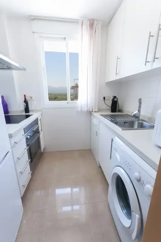 A narrow white kitchen with a window, countertop, and washing machine.