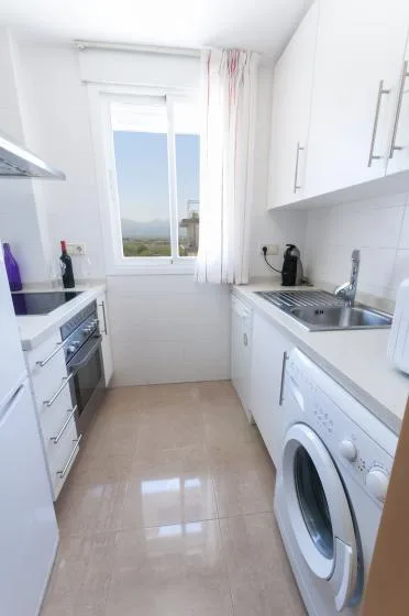 A narrow white kitchen with a window, countertop, sink, and washing machine.