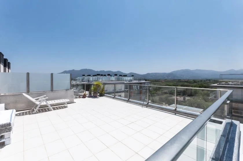 Una terraza soleada en la azotea, con suelo de baldosas claras y barandillas de vidrio, mira hacia colinas lejanas.